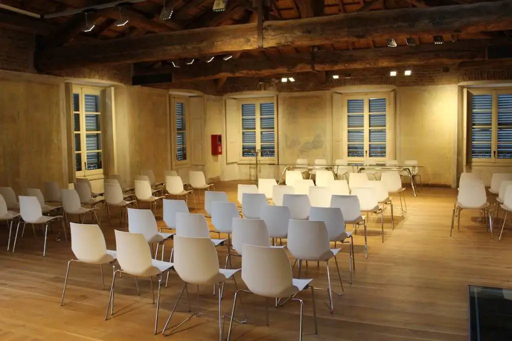 Une salle de séminaire bien éclairée avec des rangées de chaises blanches disposées pour un événement. La pièce dégage un charme rustique avec des poutres en bois apparentes, un parquet en bois et des fenêtres vintage qui créent une ambiance chaleureuse, parfaite pour réserver une salle de séminaire.